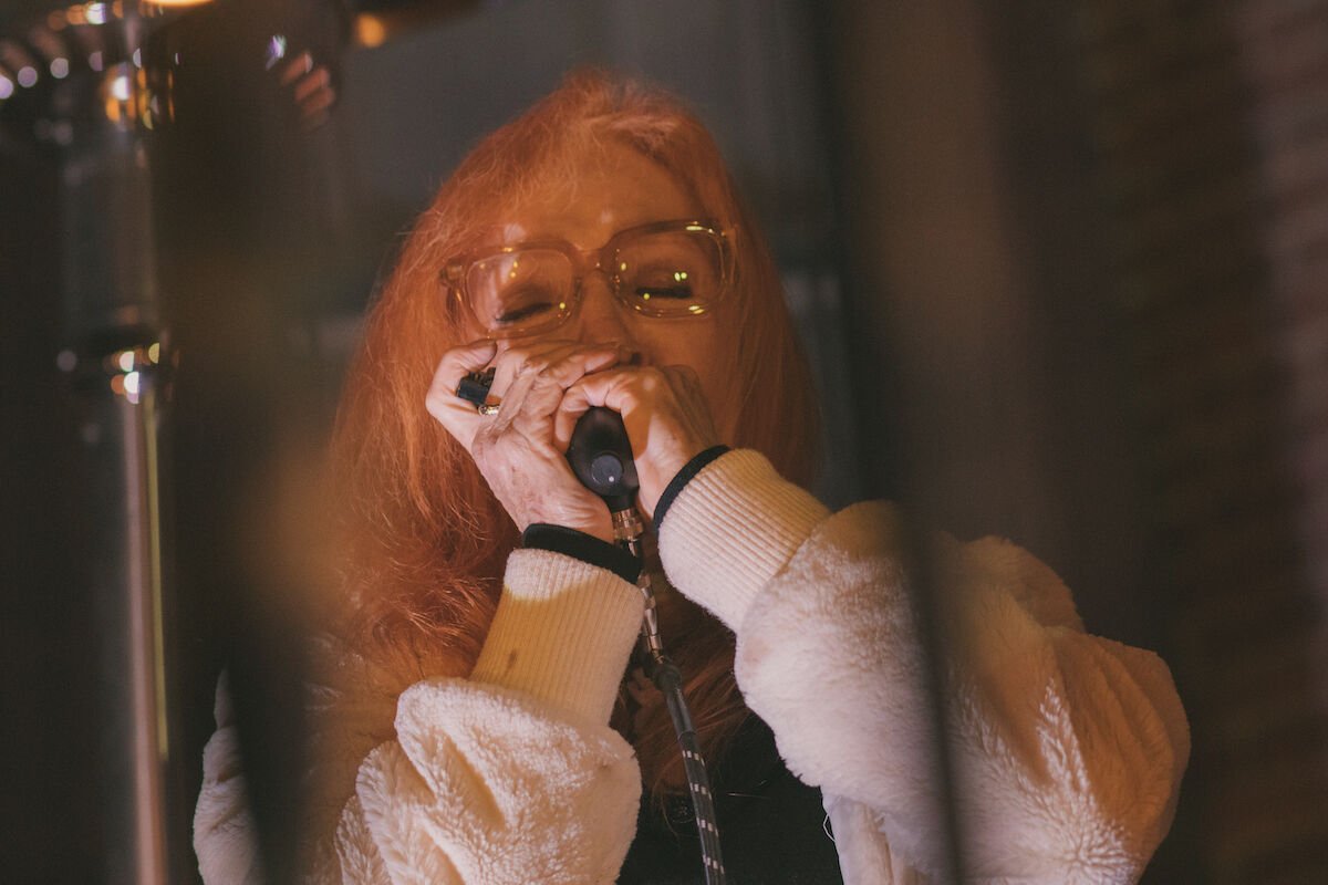 A performer with red hair and eyeglasses plays her harmonica. She's wearing a warm white, fuzzy jacket. A heating lamp keeps her warm in the background.