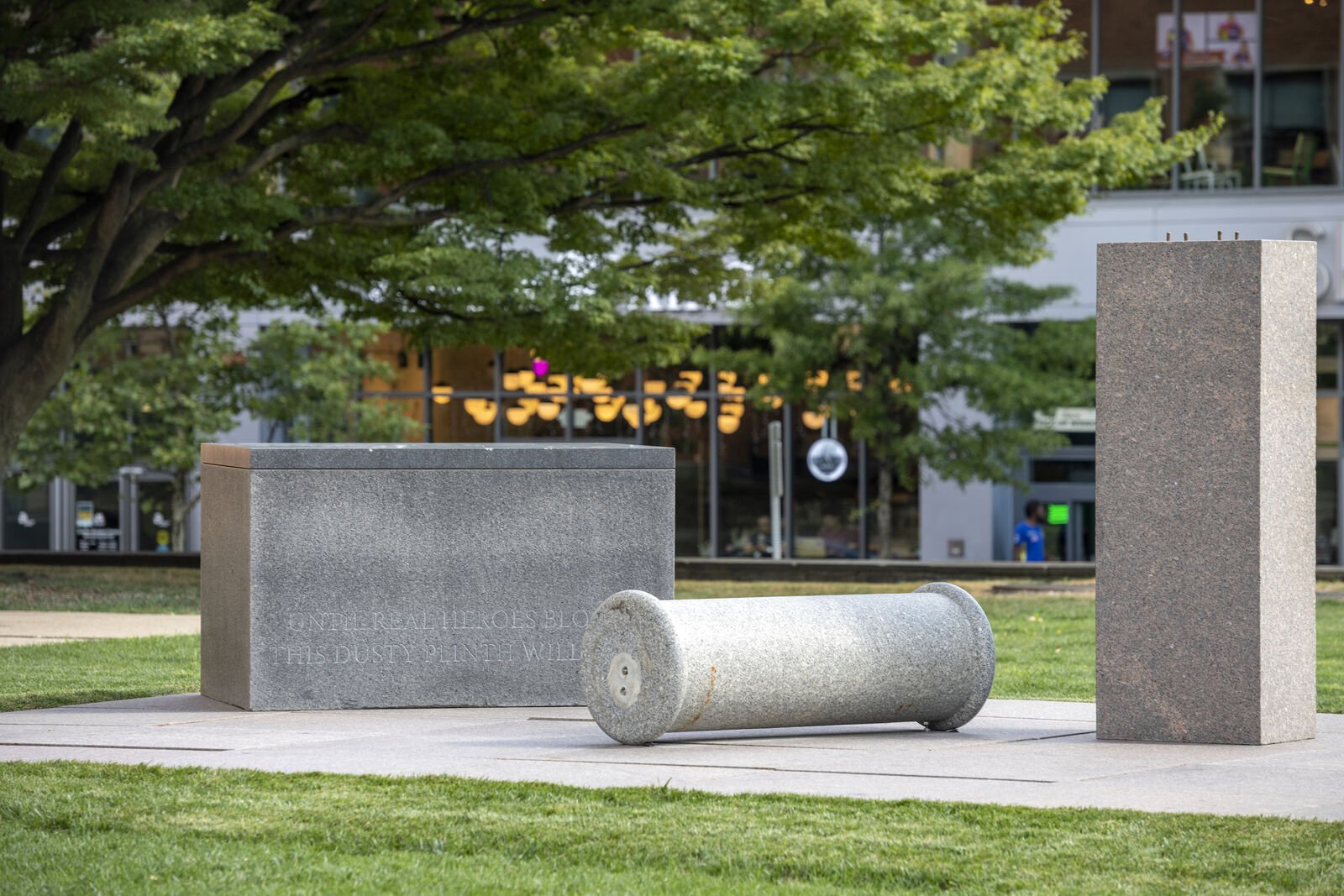 In the center, three sculptural objects in this order: a stone rectangular plint, a fallen column with rounded ends, and another column standing tall. All three are various shades of gray and rest on a base of concrete surrounded by green grass. In the background on the left, a tall tree and its branches fan out to the right.