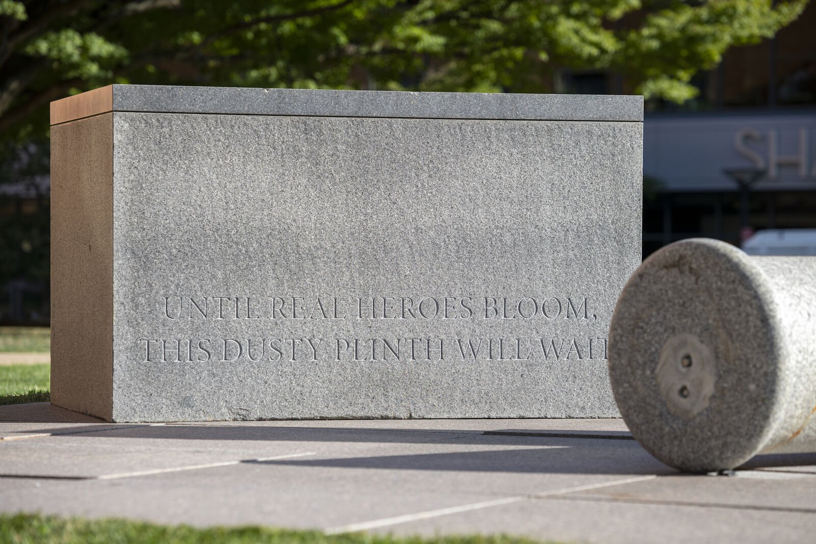 Two grey stone plinths. One in the background has an inscription that reads: Until real heroes bloom, this dusty plinth will wait. In the foreground a rounded column lays on its side.